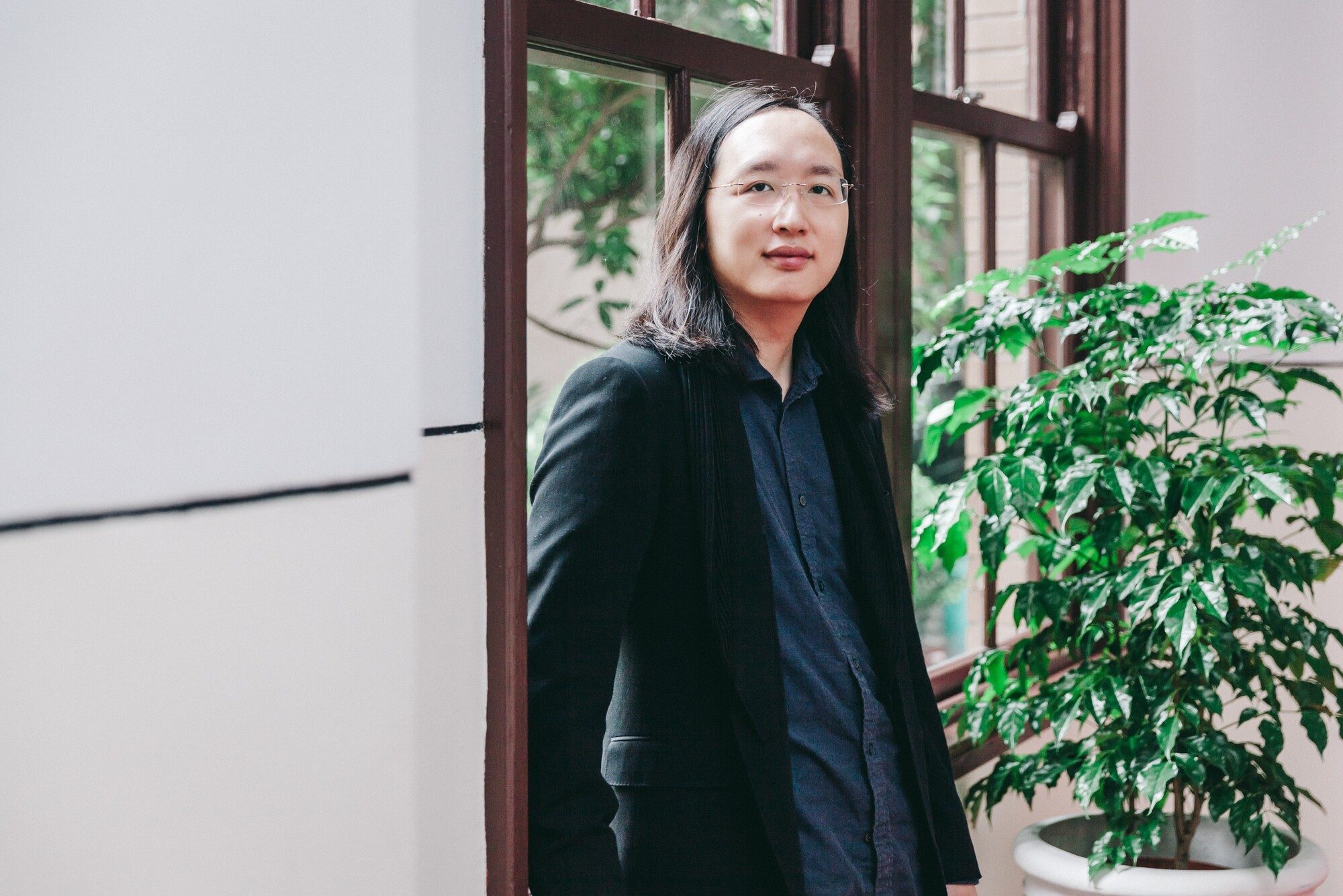 Audrey Tang in a dark blazer standing by a wood-framed window, greenery visible outside, with a calm gentle expression