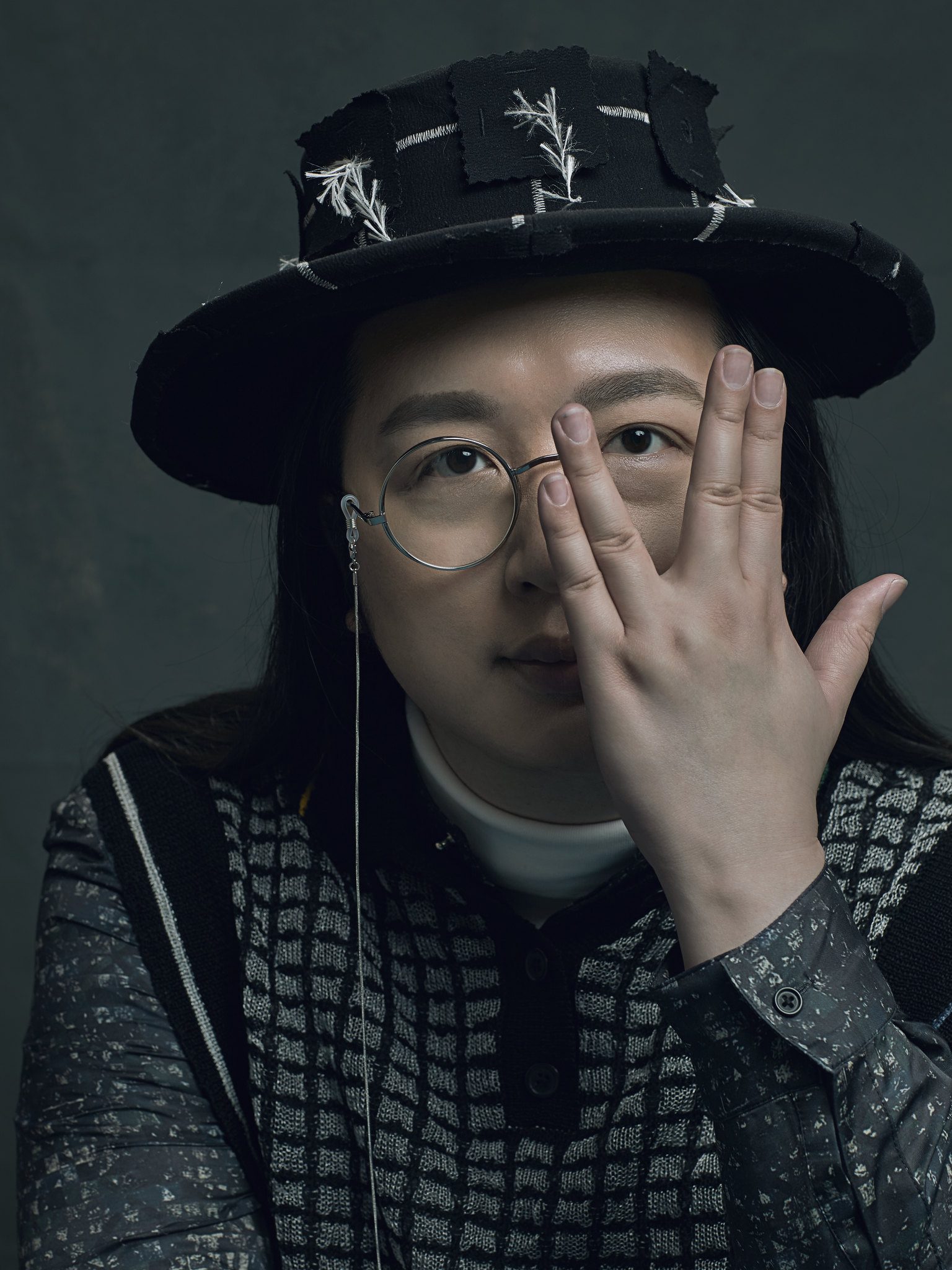 Audrey Tang in round glasses and a patchwork hat, one hand raised over their left eye, lit by moody studio light